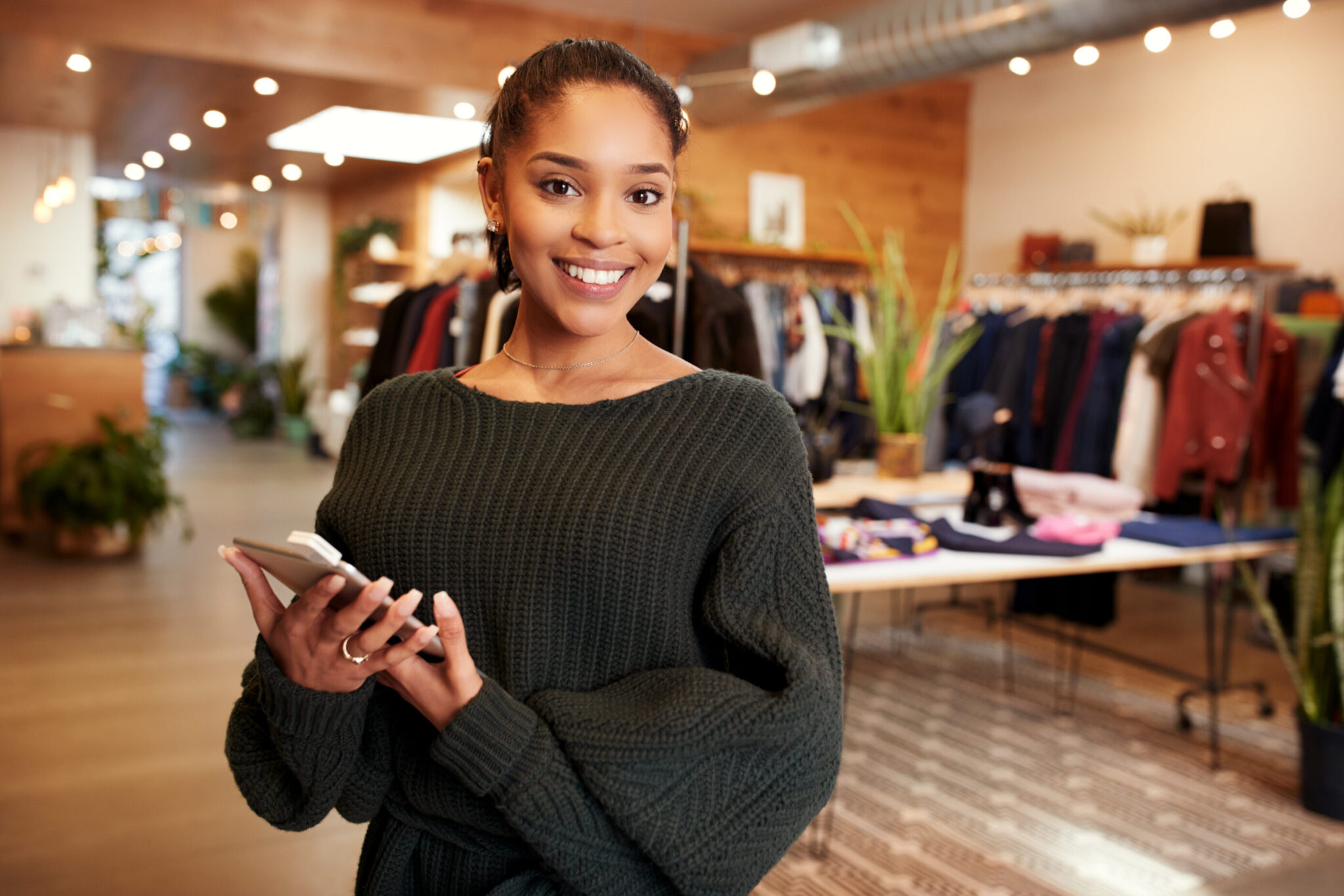 A woman smiling in front of a clothing store, possibly her own, with a tablet in hand and ready to welcome customers.